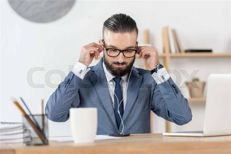 Portrait of businessman putting earphones into ears at workplace in office, stock photo