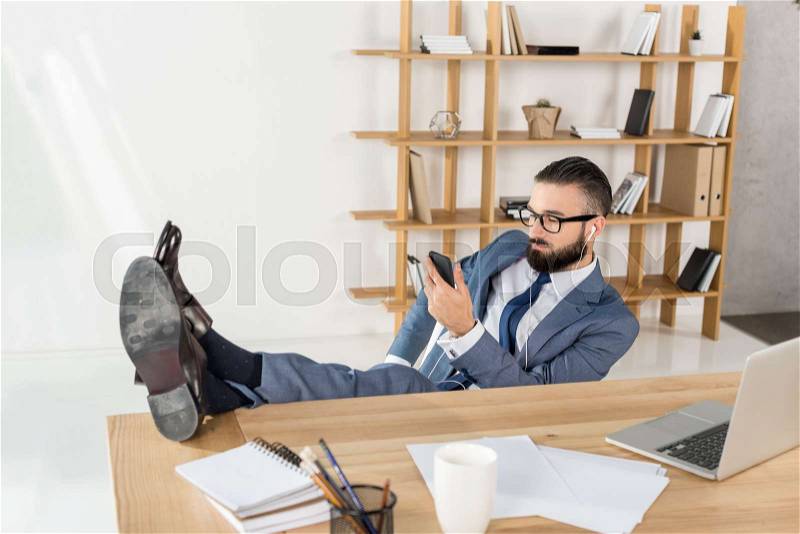 Businessman in earphones using smartphone and listening music at workplace, stock photo