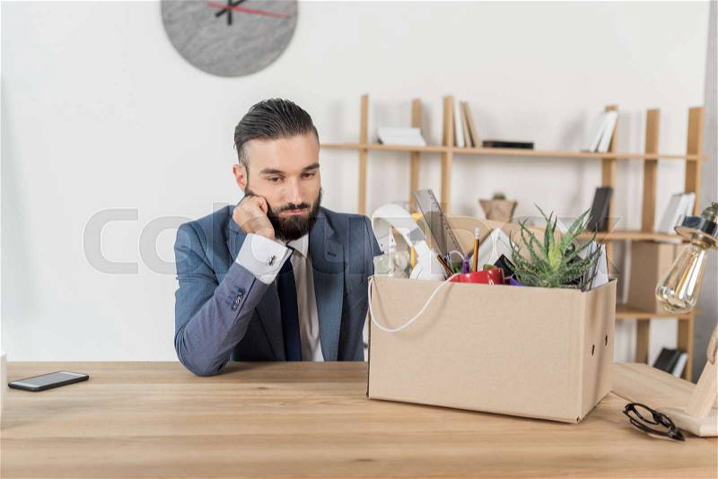 Portrait of fired upset businessman sitting at workplace with cardboard box with office supplies, stock photo