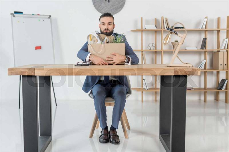 Fired upset businessman sitting at workplace with cardboard box with office supplies, stock photo