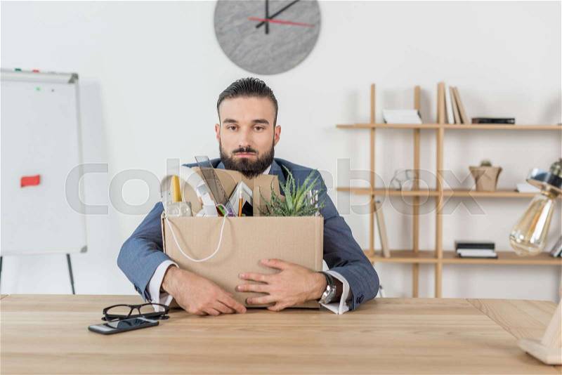 Portrait of fired upset businessman sitting at workplace with cardboard box with office supplies, stock photo