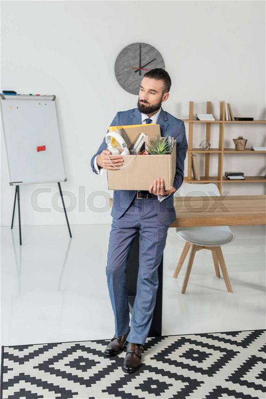 Fired sad businessman with cardboard box in hands standing at workplace in office, stock photo