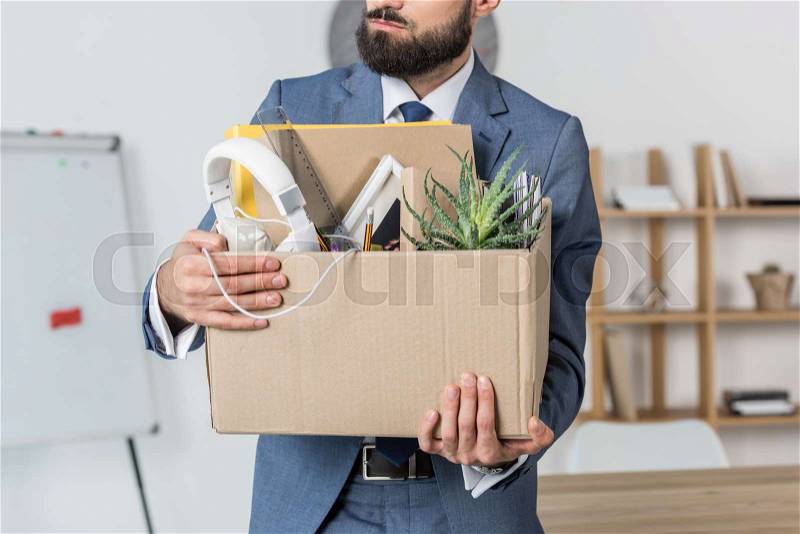 Partial view of fired businessman holding cardboard box with office supplies in office, stock photo