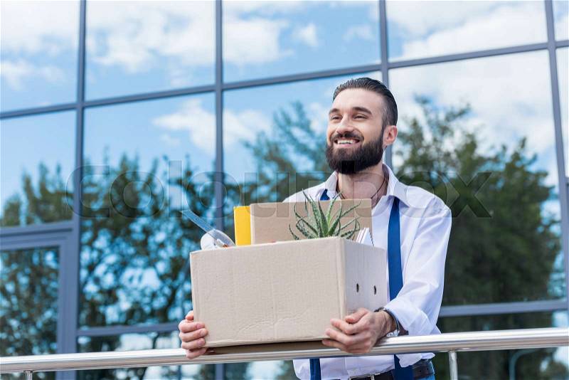 Happy businessman with cardboard box with office supplies in hands standing outside office building, quitting job concept, stock photo