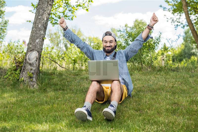 Excited man looking at laptop screen while sitting on green grass in park, stock photo