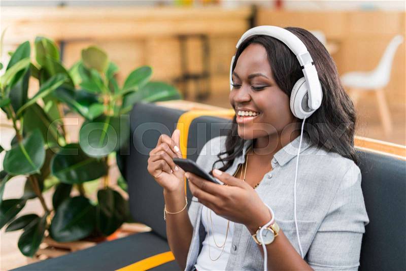 Happy african american woman listening music with headphones and smartphone while sitting in cafe, stock photo