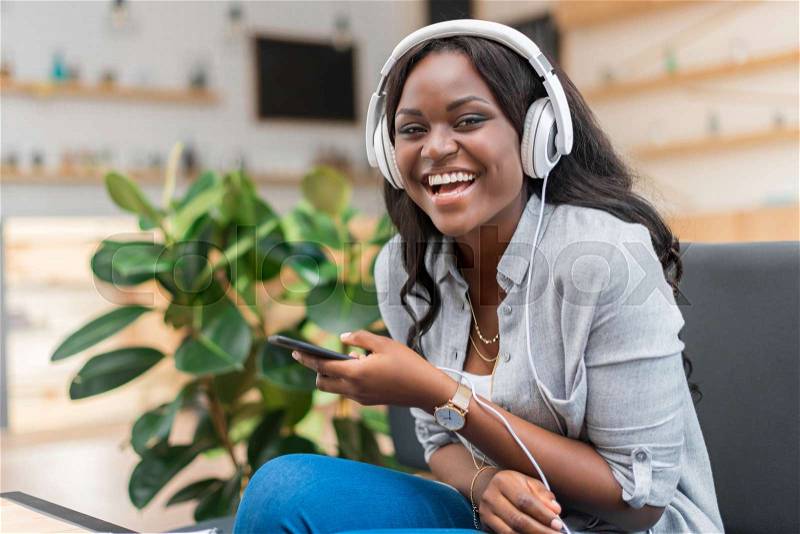 Cheerful african american woman listening music with headphones and smartphone while sitting in cafe, stock photo