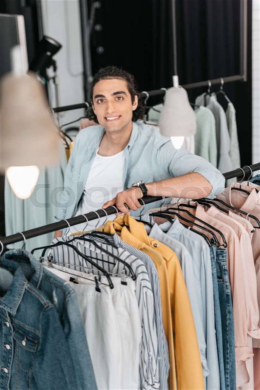 Handsome young boutique owner leaning at hangers with clothes and smiling at camera , stock photo