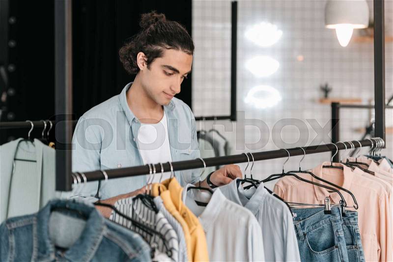 Handsome young boutique owner choosing shirts on hangers and smiling, stock photo