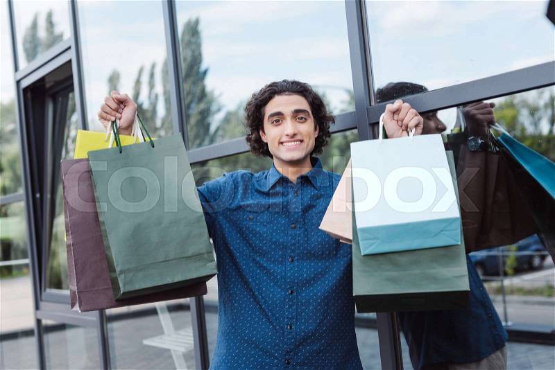 Cheerful handsome young man holding shopping bags and smiling at camera, stock photo