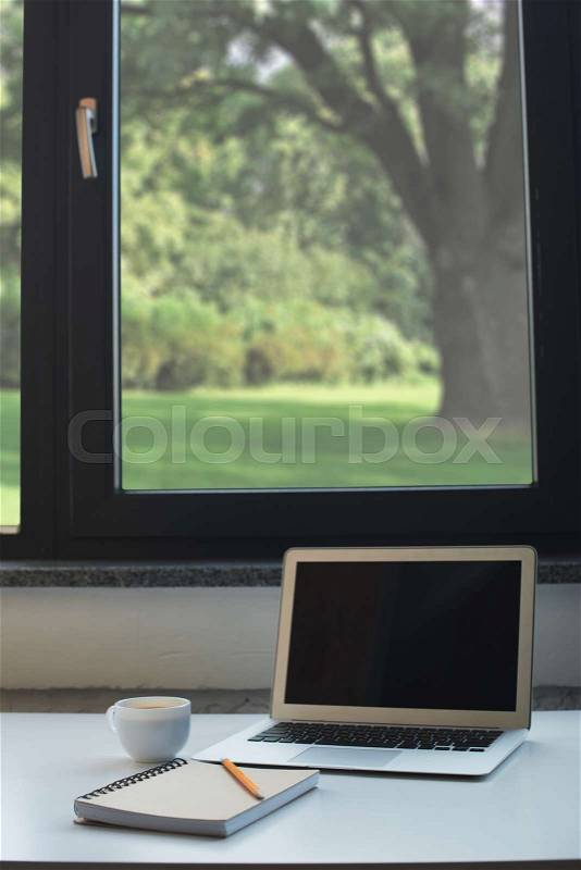 Laptop computer, notepad with pencil and cup of coffee on white desk in front of window at home office, stock photo