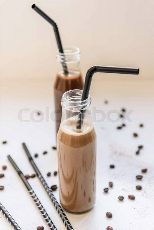 Two glass bottles with cold coffee cocktails and straws, coffee beans and cocoa powder on white table, stock photo