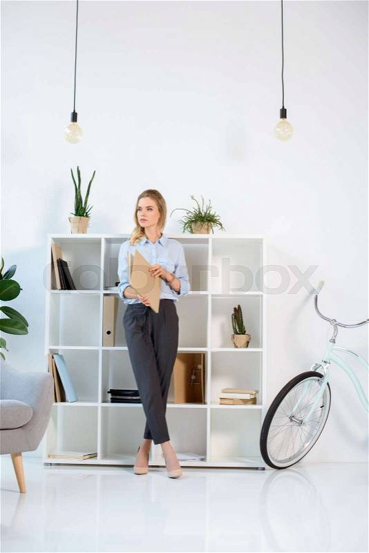 Pensive businesswoman looking away while standing with work journal in office, stock photo