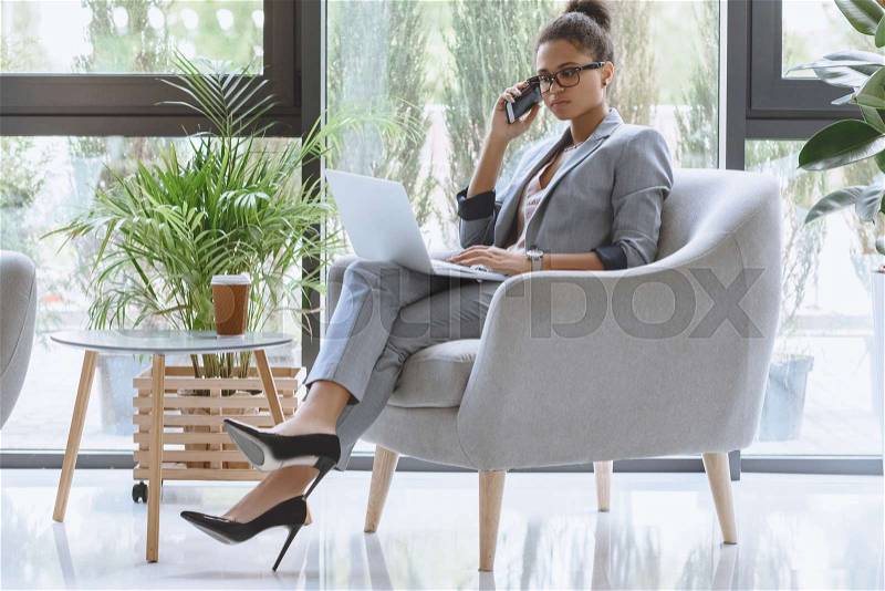 African american businesswoman sitting in armchair and talking on smartphone while working on laptop , stock photo
