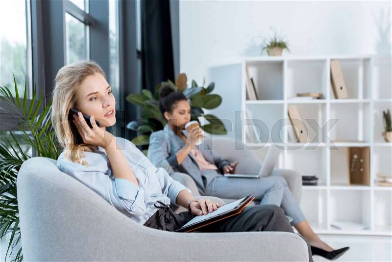 Businesswoman talking on smartphone while african american colleague drinking coffee working on laptop near by , stock photo