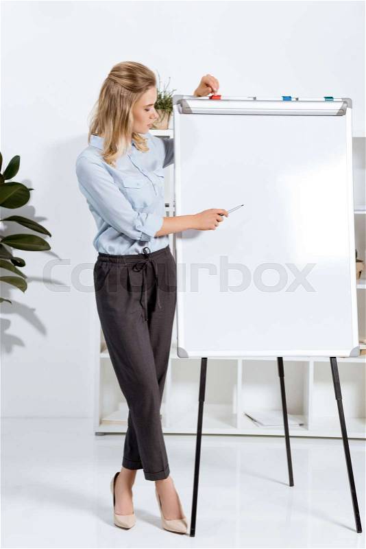 Businesswoman looking and pointing at empty white board in office, stock photo