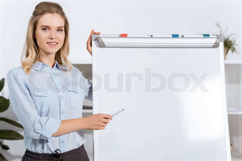 Portrait of smiling businesswoman pointing at empty white board in office, stock photo