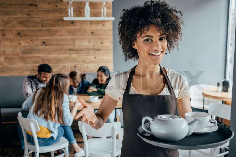 Smiling african american waitress holding tray with tea and customers sitting behind her in cafe, stock photo