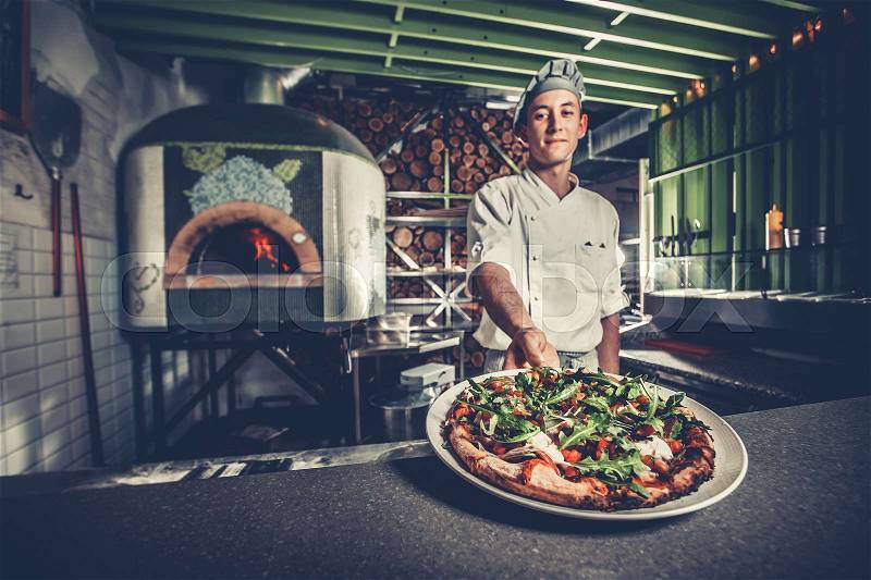 Food concept. Preparing traditional italian pizza. Young smiling chef in white uniform and gray hat show ready dish with green rucola herbs in interior of modern restaurant kitchen. Ready to eat, stock photo