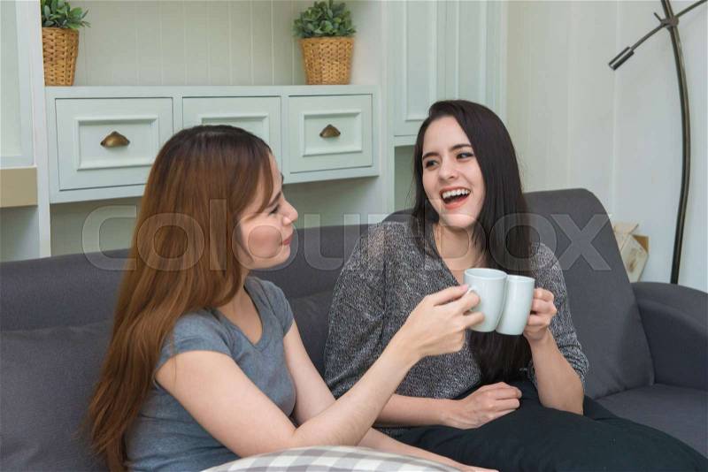Beautiful girls with friend talking and holding coffee cups in living room at home, stock photo