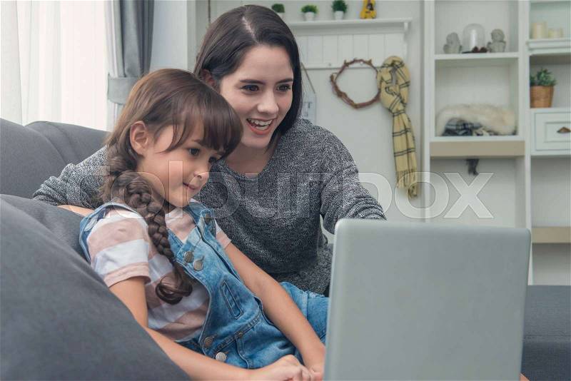 Beautiful young mom and daughter are using notebook on sofa at home, stock photo