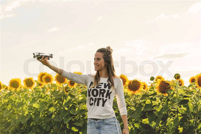 Young woman holding on hand flying mini drone outdoors. Beautiful woman is standing near a field of sunflowers at sunset, stock photo