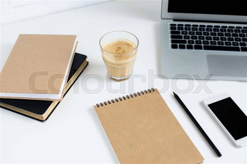 Laptop, smartphone, notepad, books and glass of coffee on white tabletop, stock photo