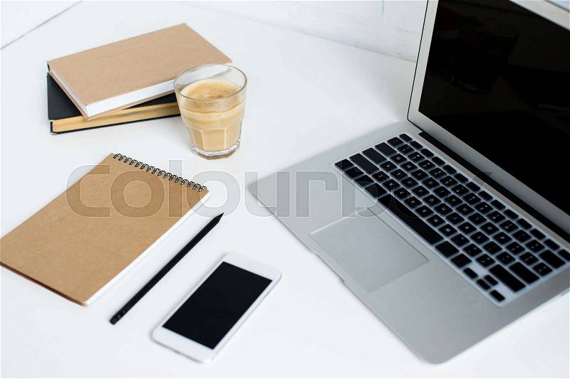 Laptop, smartphone, notepad, books and glass of coffee on white tabletop, stock photo