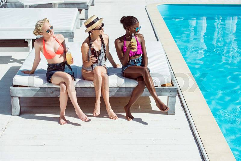 Young beautiful multiethnic women resting with beverages at poolside at resort, stock photo