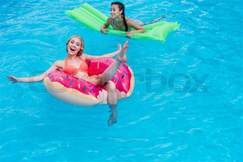 Young multiethnic women floating on inflatable mattresses in swimming pool at resort, stock photo