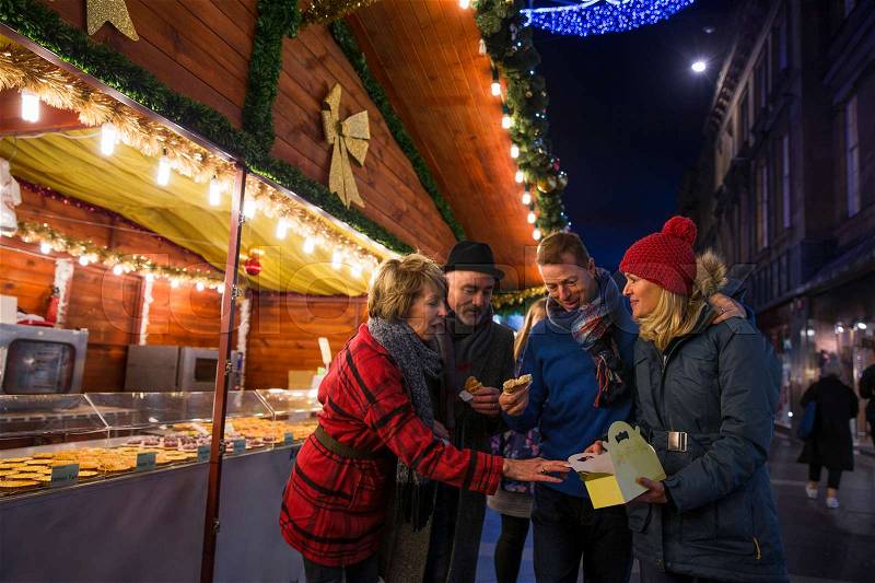 Mature friends are at a christmas market in the city. They are sharing some festive pastries that they have bought from one of the stalls, stock photo