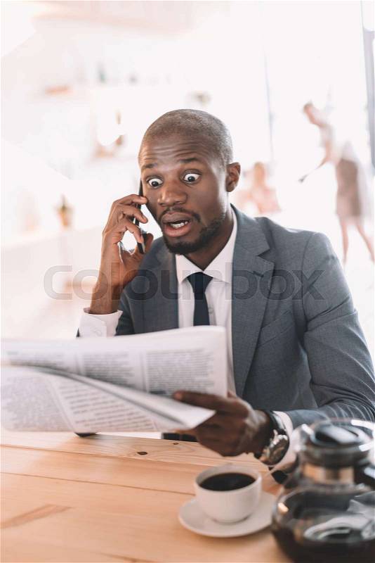 Shocked handsome african american businessman with smartphone reading newspaper during coffee break in cafe , stock photo