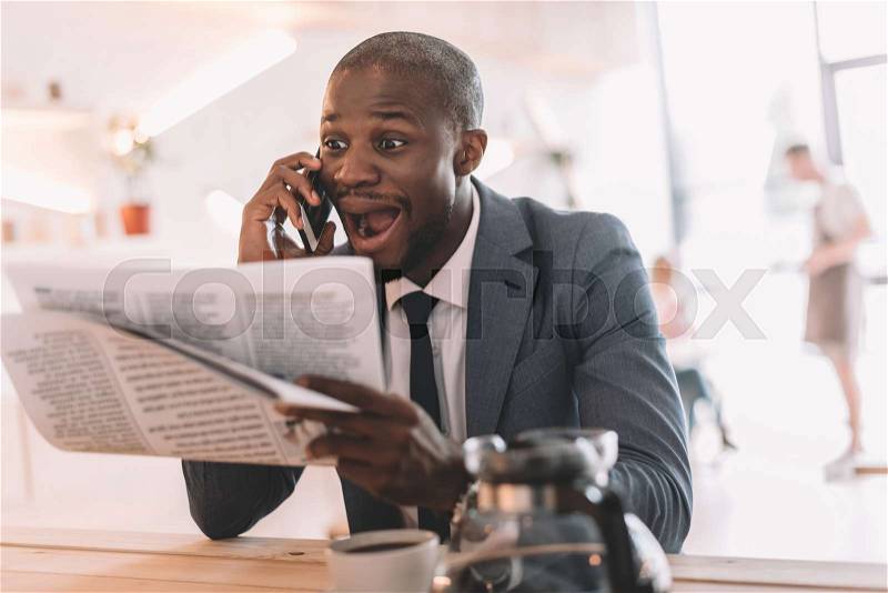 Excited handsome african american businessman with smartphone reading newspaper during coffee break in cafe , stock photo