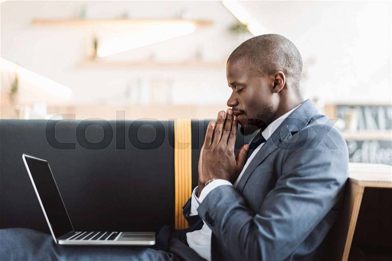 Smiling african american businessman with closed eyes praying with laptop, stock photo