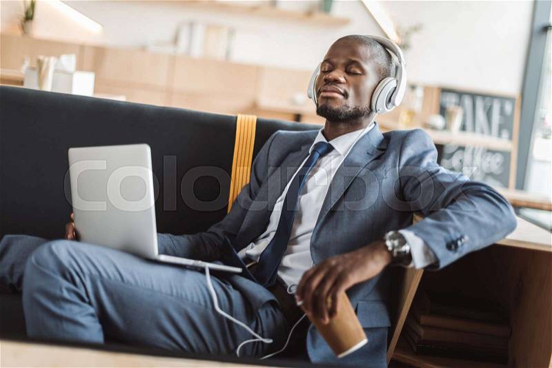 African american businessman sitting with disposable cup of coffee and listening music with headphones and laptop in cafe , stock photo