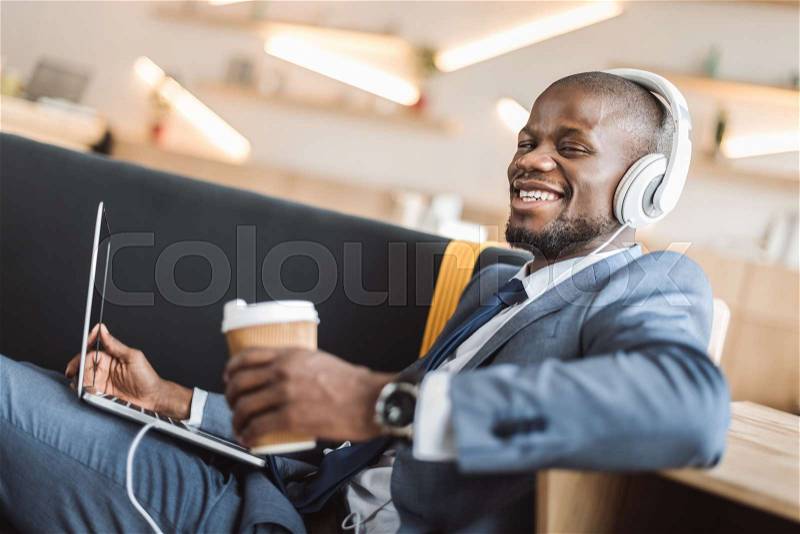 African american businessman sitting with disposable cup of coffee and listening music with headphones and laptop in cafe , stock photo