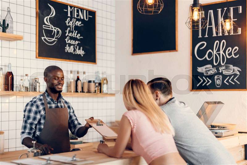 Handsome smiling african american barista giving menu to clients on bar counter in cafe , stock photo