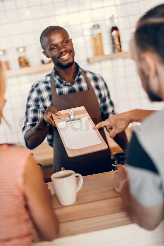 Handsome smiling african american barista giving menu to clients on bar counter in cafe , stock photo
