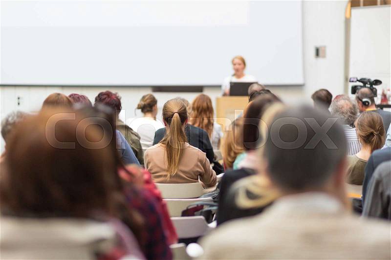 Female speaker giving presentation in lecture hall at university workshop. Audience in conference hall. Rear view of unrecognized participant in audience. Scientific conference event, stock photo
