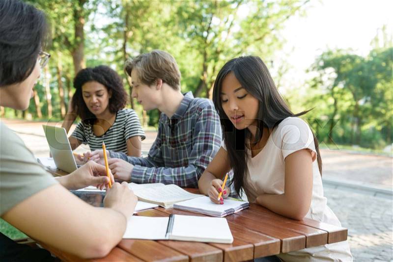 Picture of multiethnic group of young students sitting and studying outdoors while talking. Looking aside, stock photo