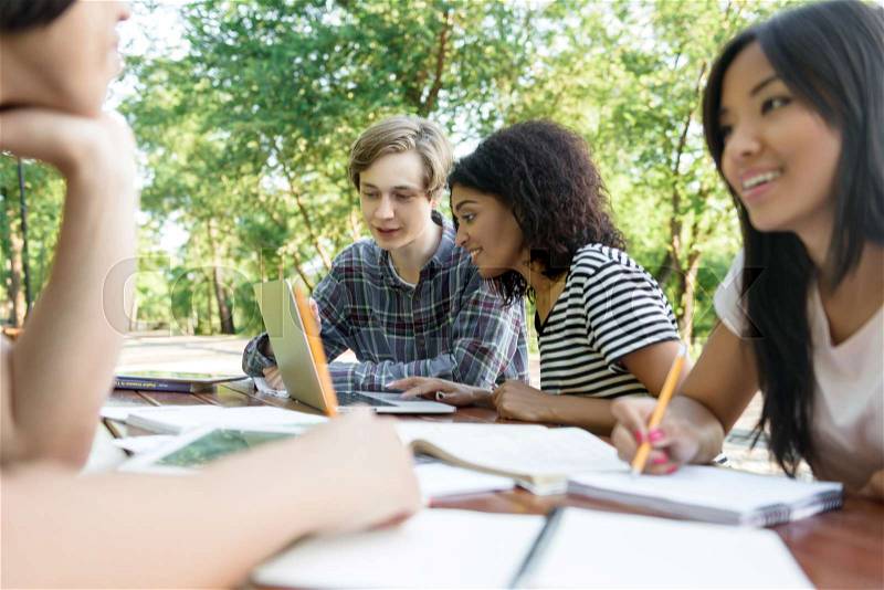 Photo of multiethnic group of young smiling students sitting and studying outdoors while talking. Looking aside, stock photo