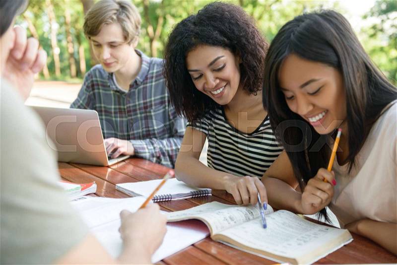 Image of multiethnic group of young happy students sitting and studying outdoors while talking. Looking aside, stock photo