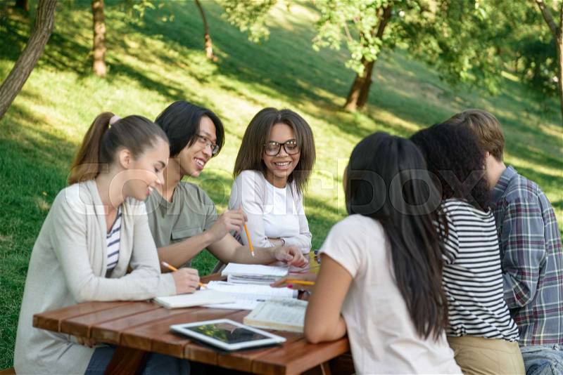 Picture of multiethnic group of young students sitting and studying outdoors while talking. Looking aside, stock photo