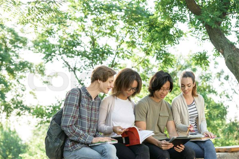 Picture of multiethnic group of young cheerful students sitting and studying outdoors. Looking aside, stock photo