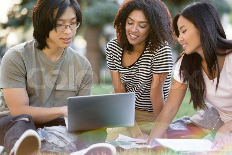 Image of young happy group of multiethnic students studying outdoors using laptop computer. Looking aside, stock photo