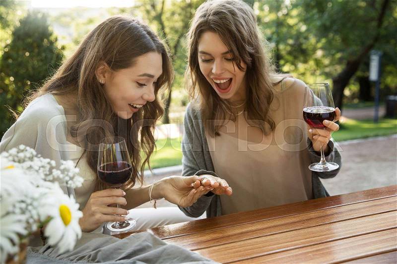 Image of happy young two women sitting outdoors in park drinking wine. Looking aside, stock photo