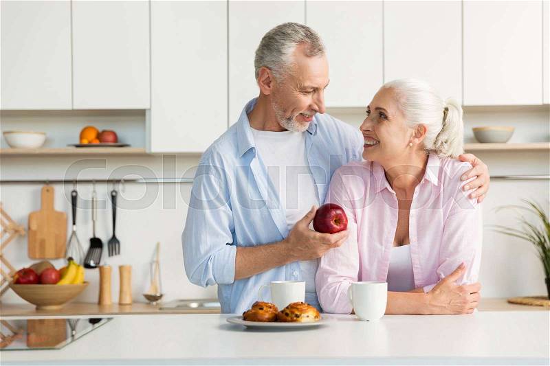 Picture of smiling mature loving couple family standing at the kitchen. Man holding apple. Looking at each other, stock photo