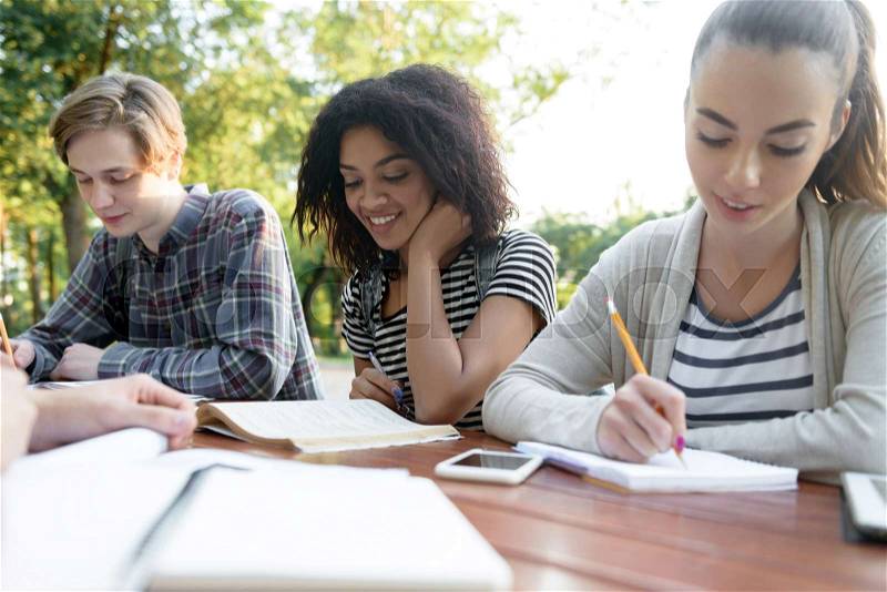 Multiethnic group of happy young people friends sitting and studying outdoors. Looking aside, stock photo