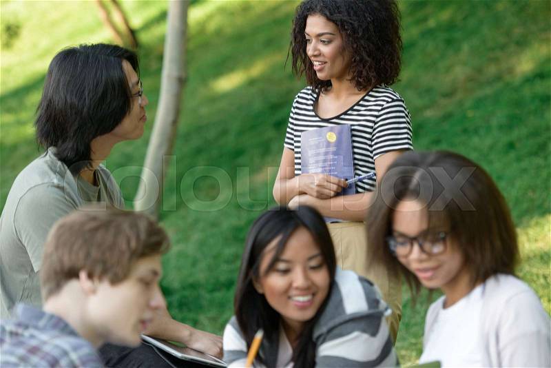 Image of multiethnic cheerful group of young students sitting and studying outdoors while using laptop. Looking aside, stock photo