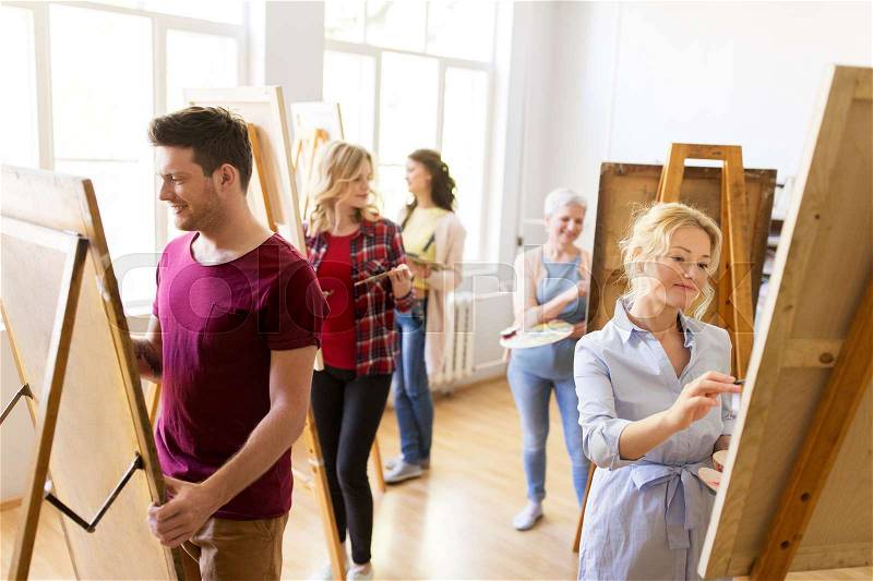 Creativity, education and people concept - group of artists or students with brushes and palettes painting on easels at art school studio, stock photo